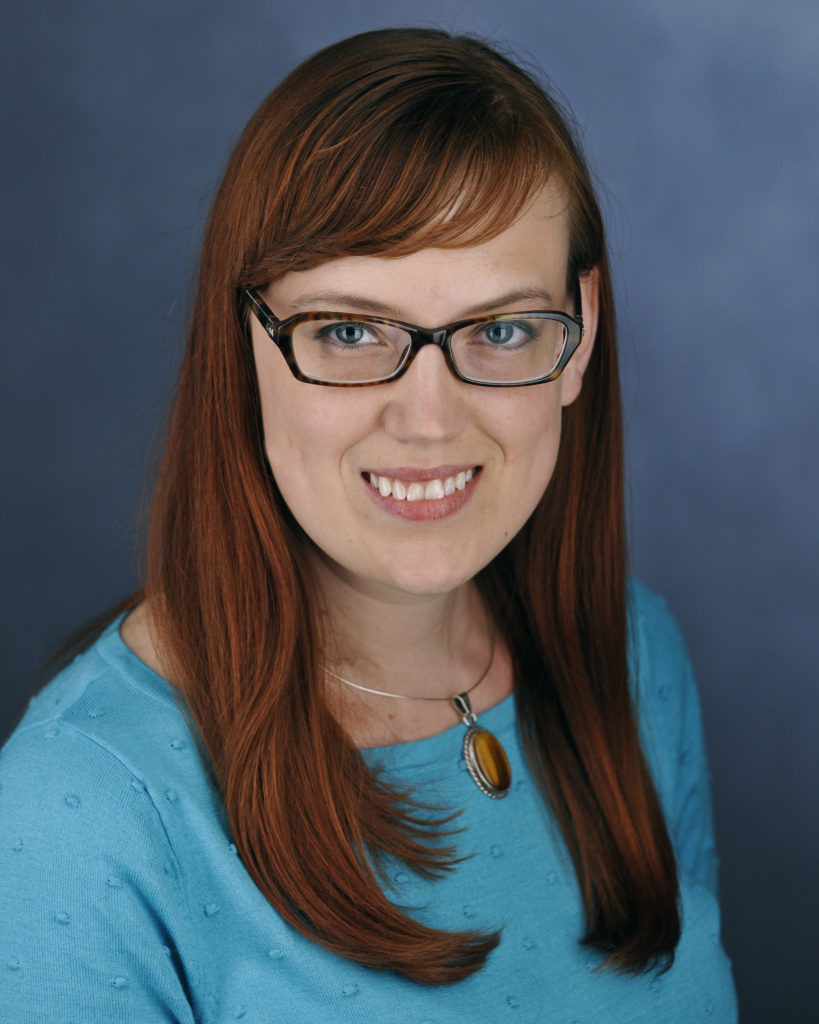 Photo of Carrie Wendell-Hummell. She is sitting in front of a blue background and smiling.