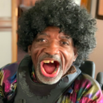 Photo of Monnie Hall sitting in a room. He is wearing a colorful shirt and smiling.